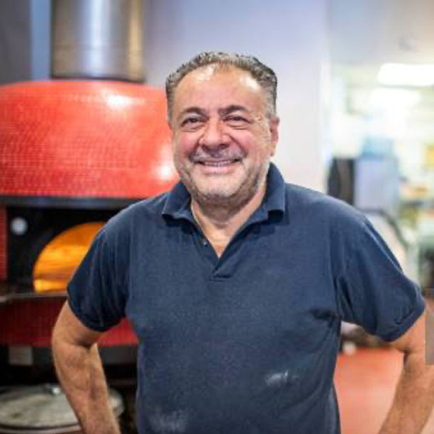Lächelnder Mann in dunkelblauem Poloshirt steht vor einem offenen Pizzaofen in einer Küche.