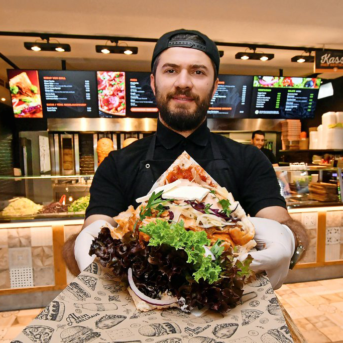 Ein Koch in schwarzer Kleidung und Handschuhen hält ein großes, gefülltes Döner-Sandwich mit Salat und Soßen in einem Schnellimbiss.
