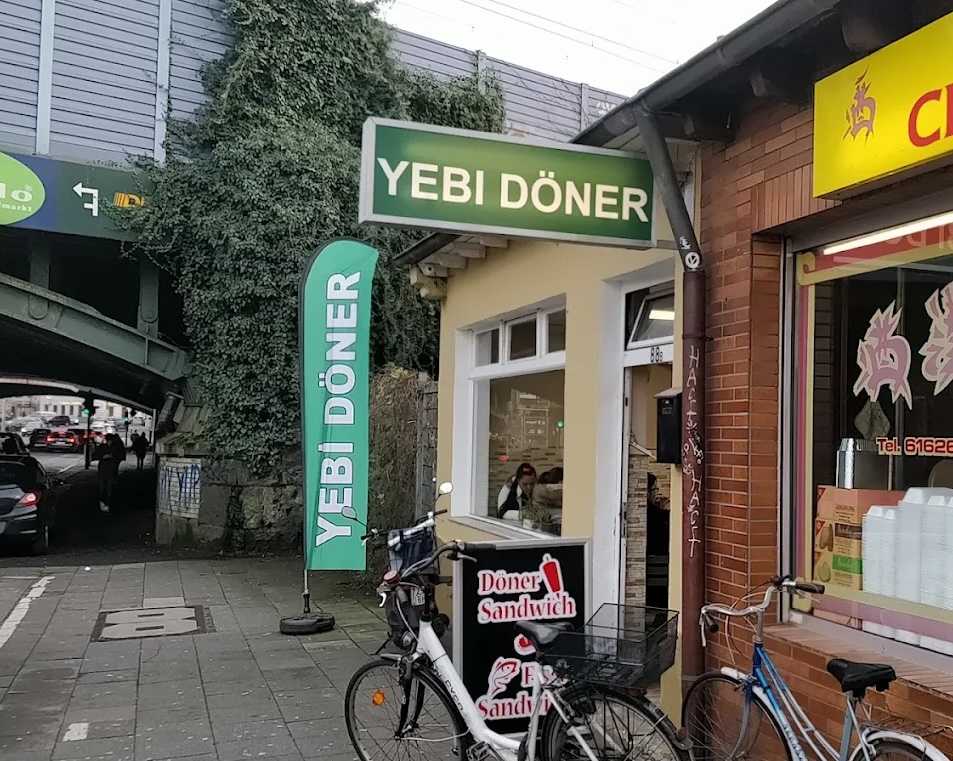Außenansicht eines Dönerladens mit grünem Schild und Fahne mit der Aufschrift 'YEBI DÖNER' und zwei Fahrrädern davor.