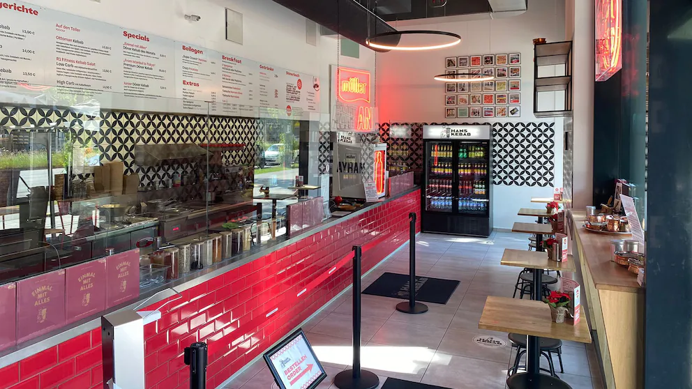 Innenansicht eines modernen Schnellrestaurants mit roten Fliesen an der Theke, Tischen mit Stühlen, Getränkekühlschrank und Speisekarte an der Wand.