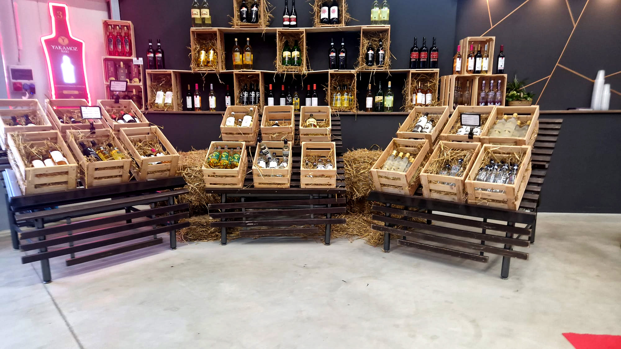 Drei Holztische mit Weinkisten gefüllt mit verschiedenen Flaschen vor dunkelgrauer Wand mit Weinfässerregal und Neonflasche.