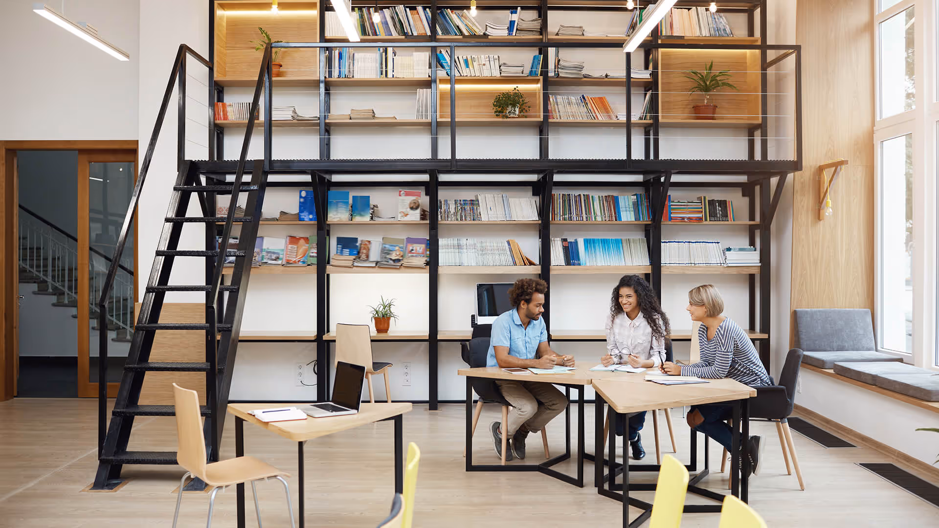Três pessoas conversando e colaborando em um escritório moderno com estantes, plantas e escada preta.
