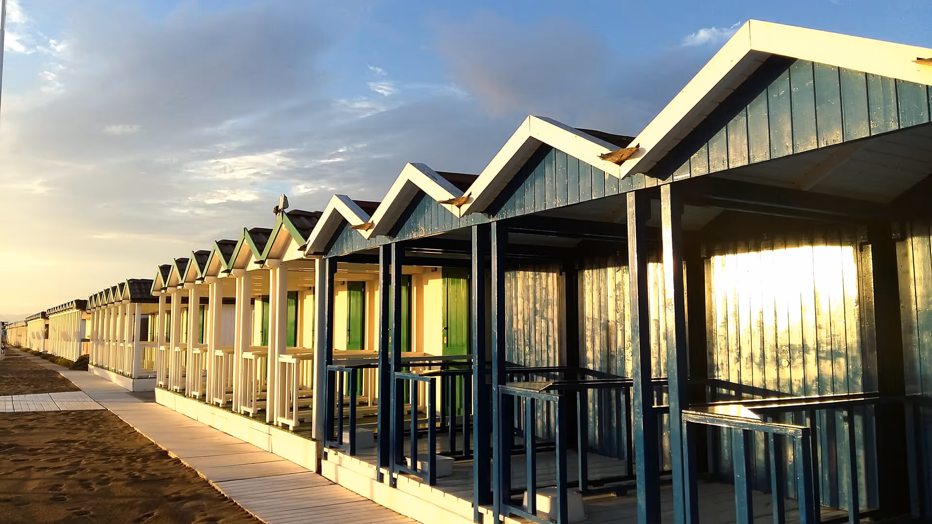 Fileiras de pequenas cabanas de madeira na praia ao entardecer com luz dourada refletindo.