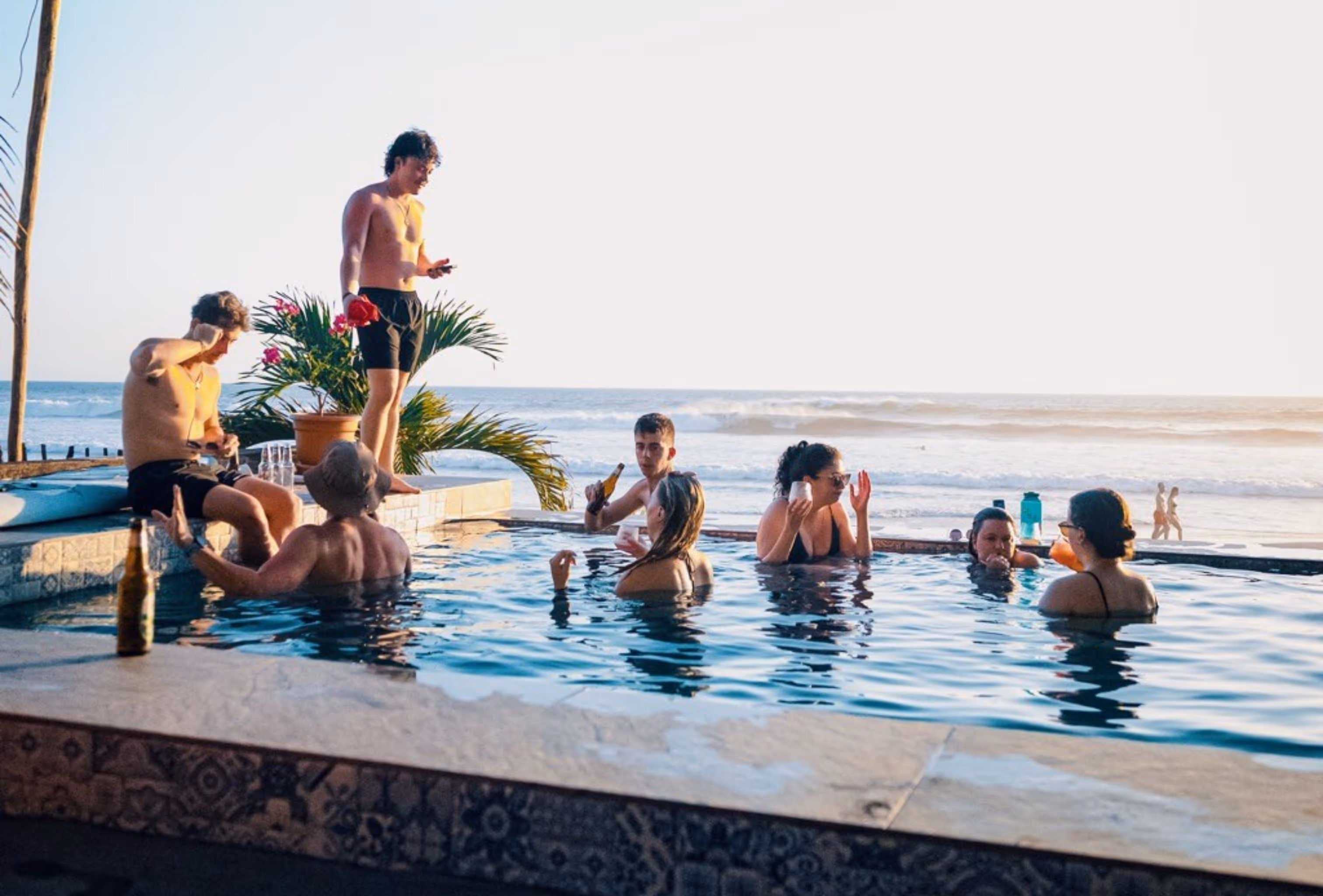 Group of people relaxing and drinking in a pool overlooking the ocean at sunset.