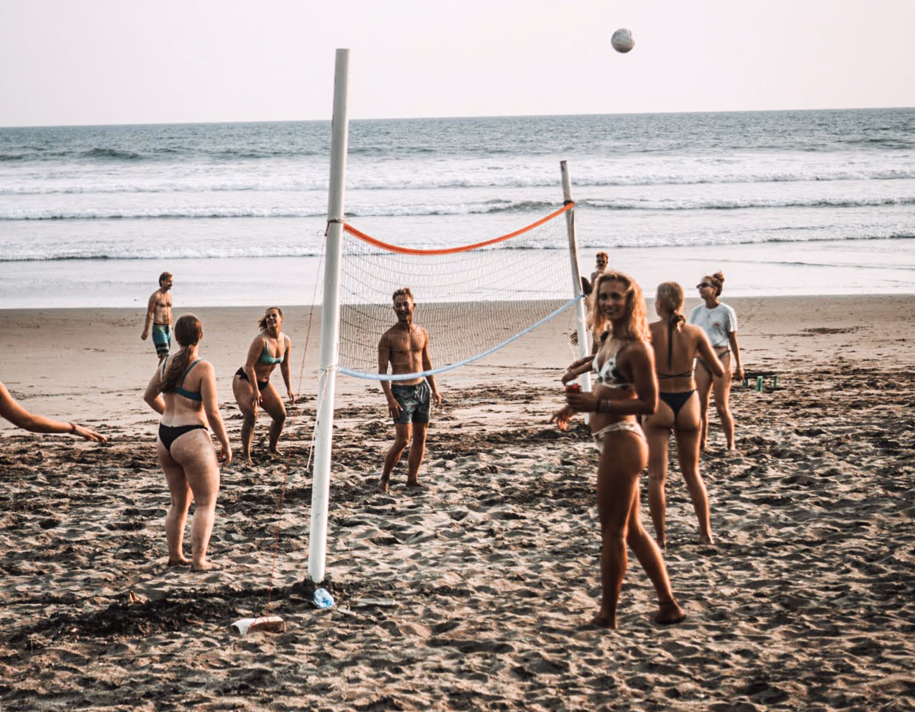 Group of people playing beach volleyball near the ocean with waves in the background.