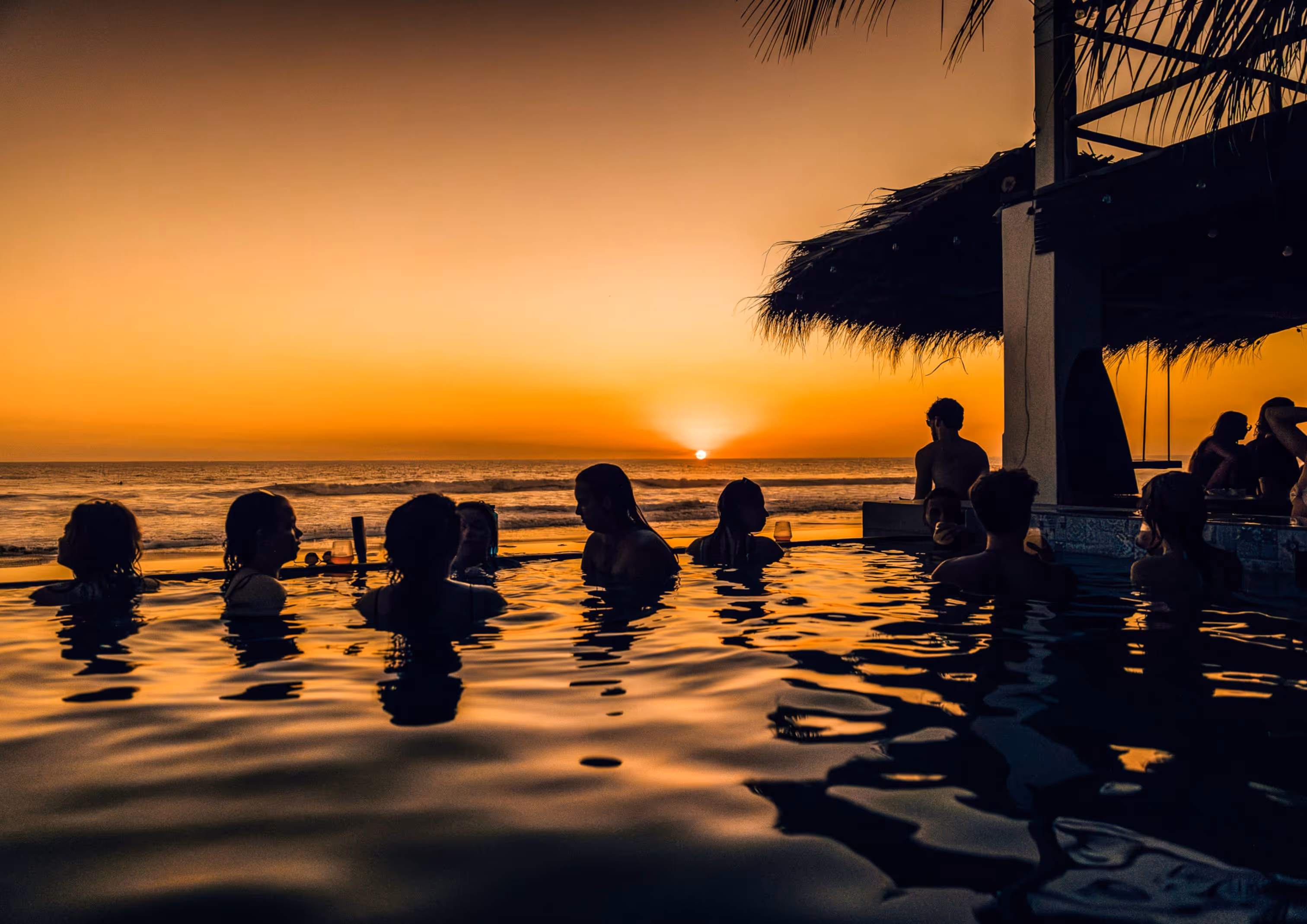 Silhouettes of people enjoying a pool at sunset by the ocean with golden sky and thatched roof structure.