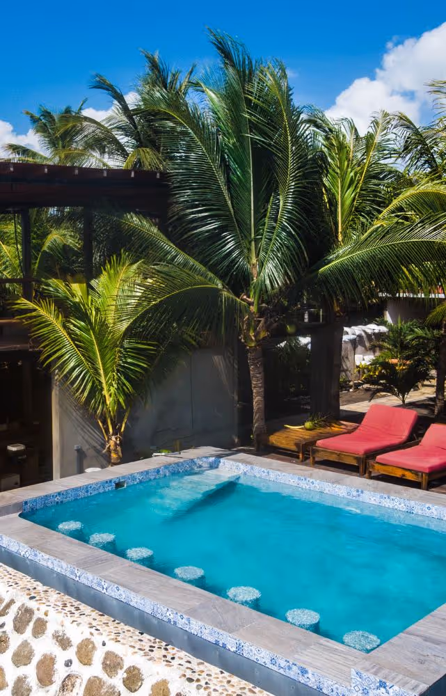 Small rectangular pool with submerged stools, surrounded by palm trees and two red lounge chairs under a clear blue sky.
