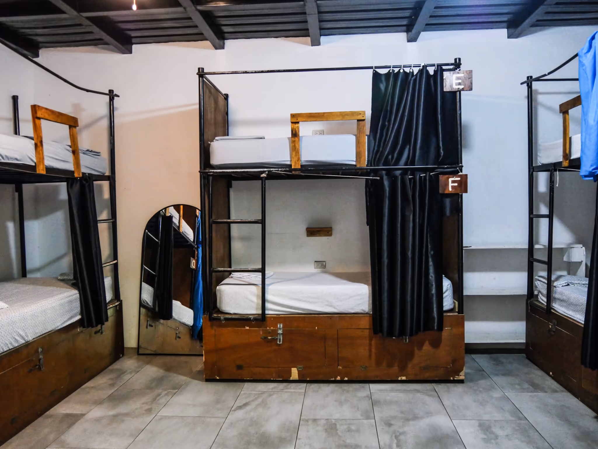 Dormitory room with wooden-framed metal bunk beds, some with privacy curtains labeled E and F, and a floor mirror leaning against the wall.
