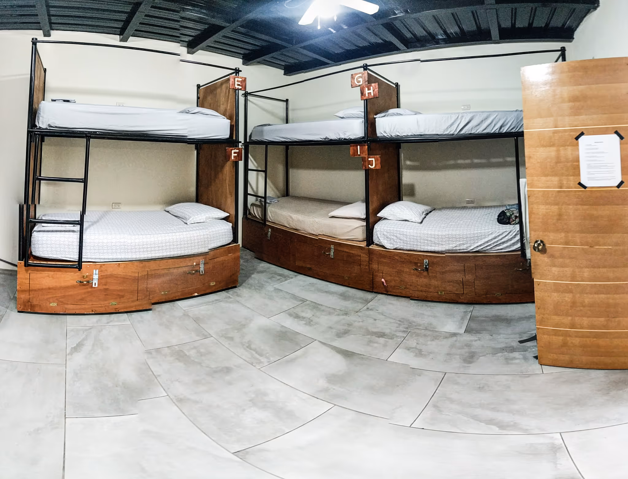 Room with three wooden bunk beds labeled E-J, white bedding, and a partially open wooden door with a paper taped on it.