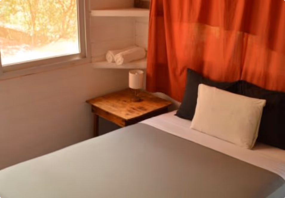 Bed with white and black pillows, gray blanket, wooden nightstand with lamp, and window with natural light.