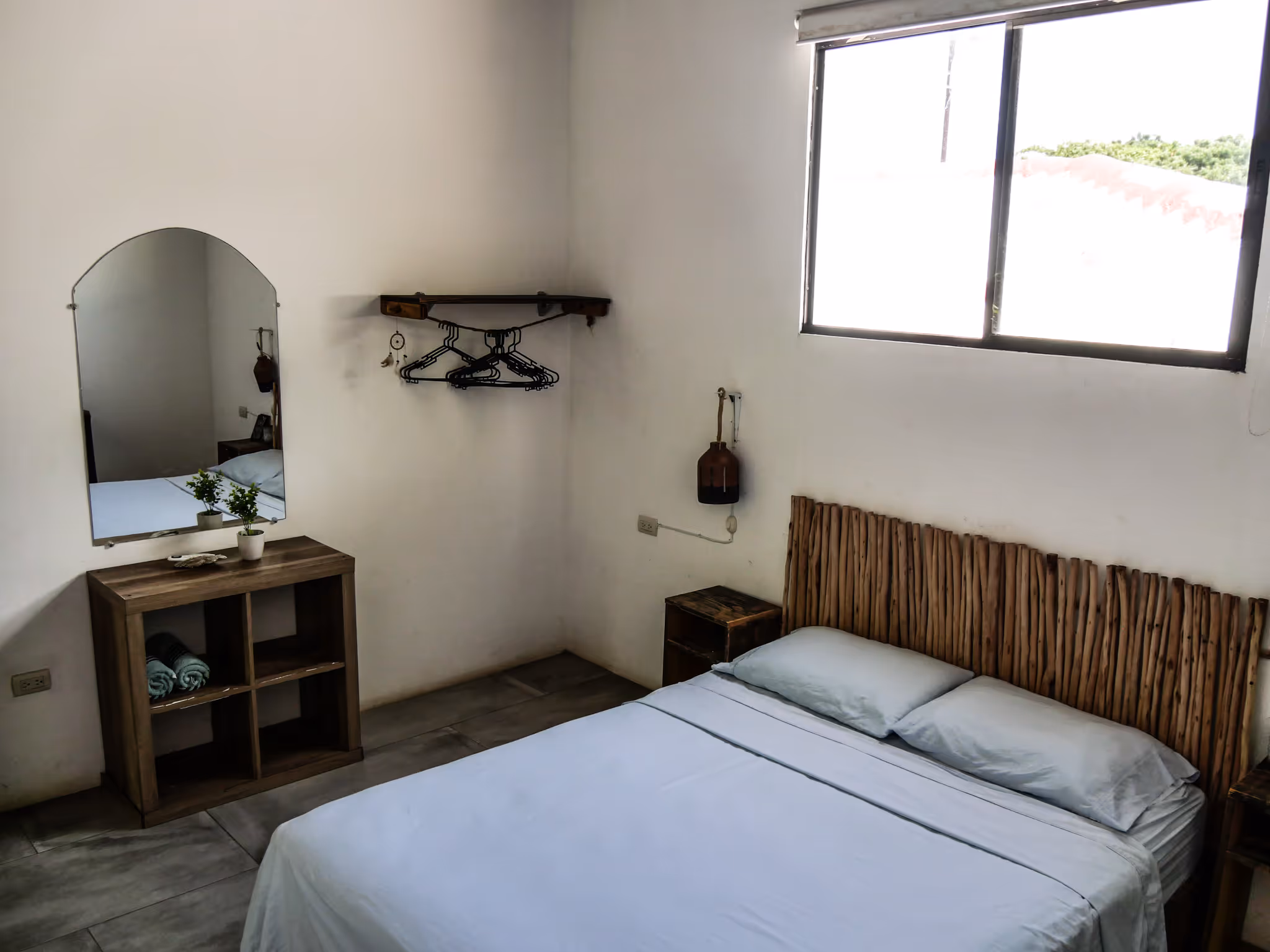 Minimalist bedroom with a wooden headboard, a bed with light blue sheets, a small wooden nightstand, a wall-mounted clothing rack with hangers, and a wooden cubby shelf with a mirror above it.