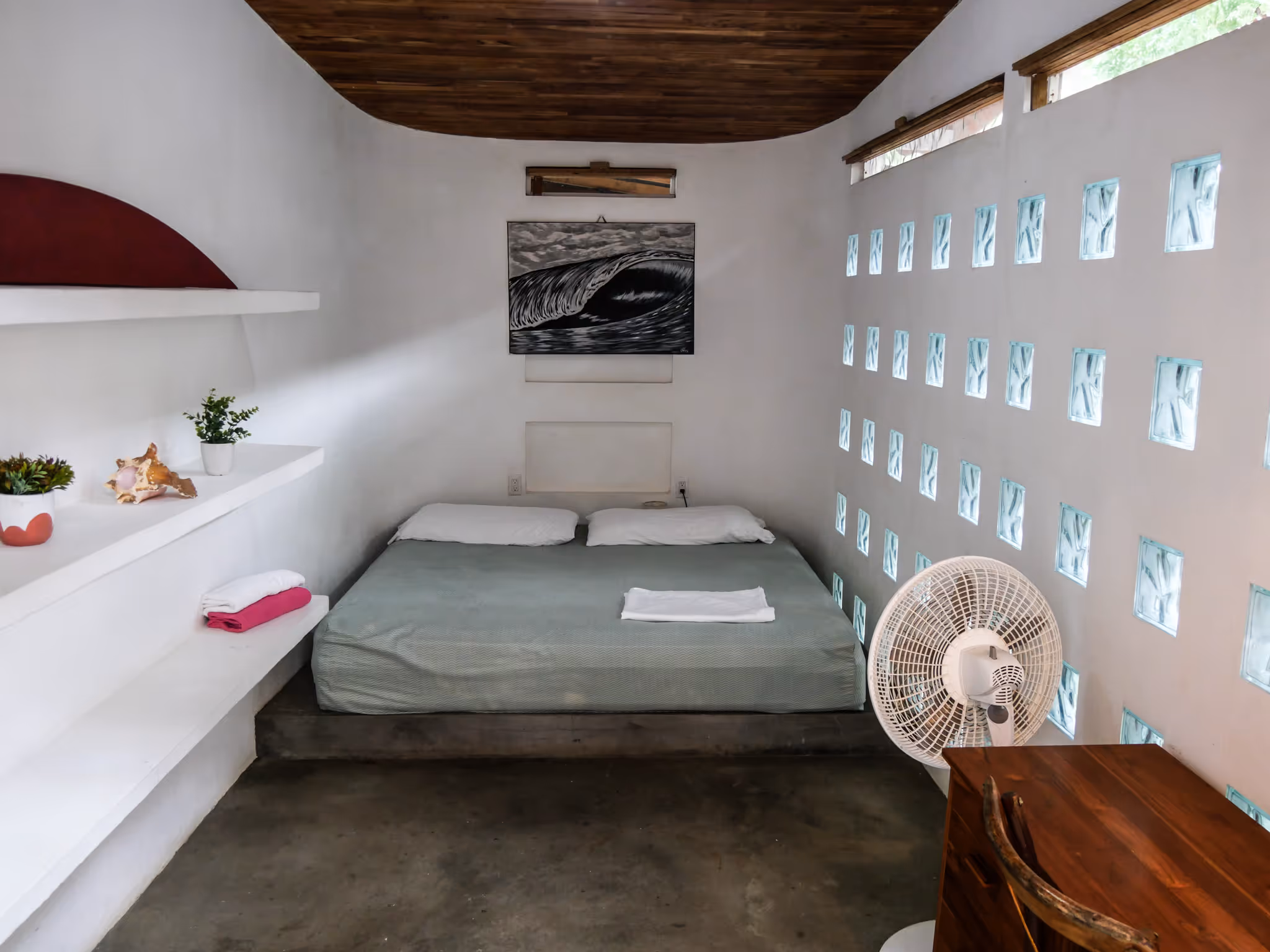 Minimalist bedroom with a low bed covered in a light green blanket, two pillows, and a folded towel, white textured walls with glass blocks letting in light, wooden ceiling, a desk with a chair, standing fan, and shelves decorated with plants and a seashell.