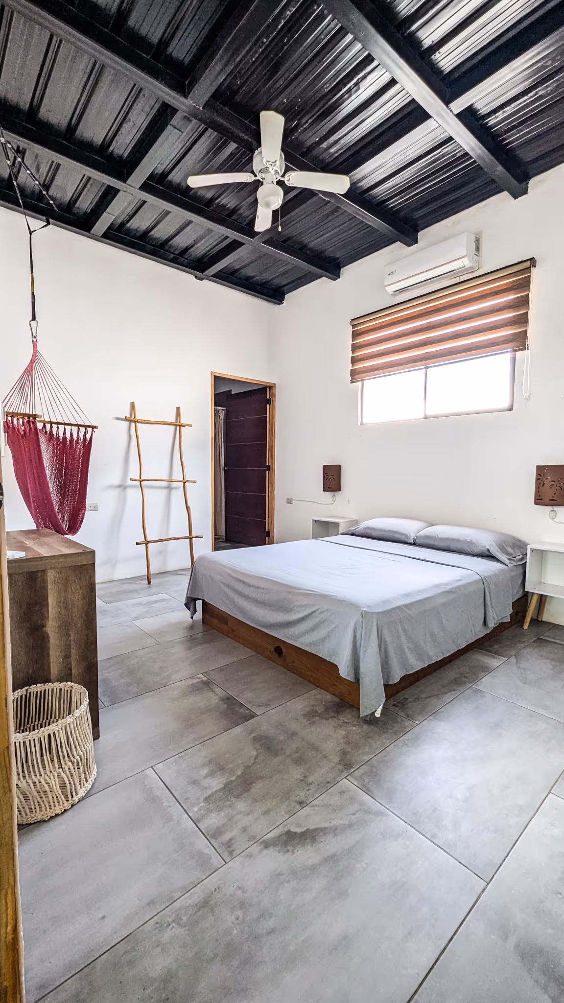 Minimalist bedroom with a gray bed, wooden furniture, a purple hanging chair, and a ceiling fan under a black metal roof.