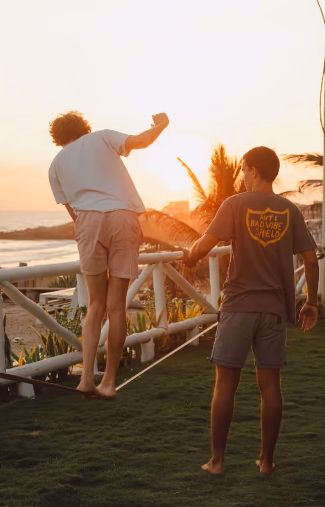 Two boys on grass near a beach at sunset, one balancing on a slackline while the other holds his hand for support.