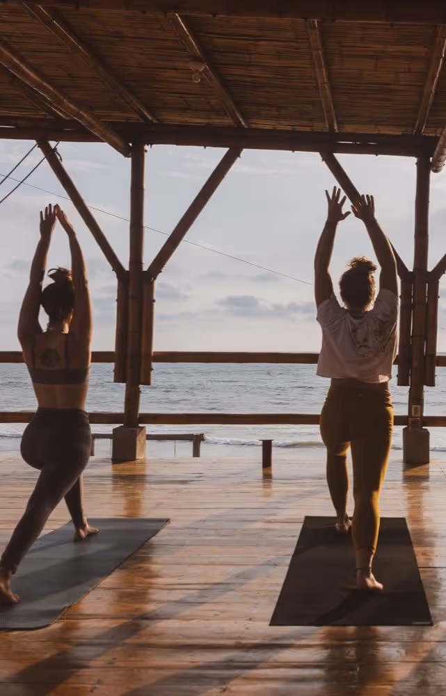 Two women practicing yoga on mats in a wooden pavilion overlooking the ocean at sunrise or sunset.