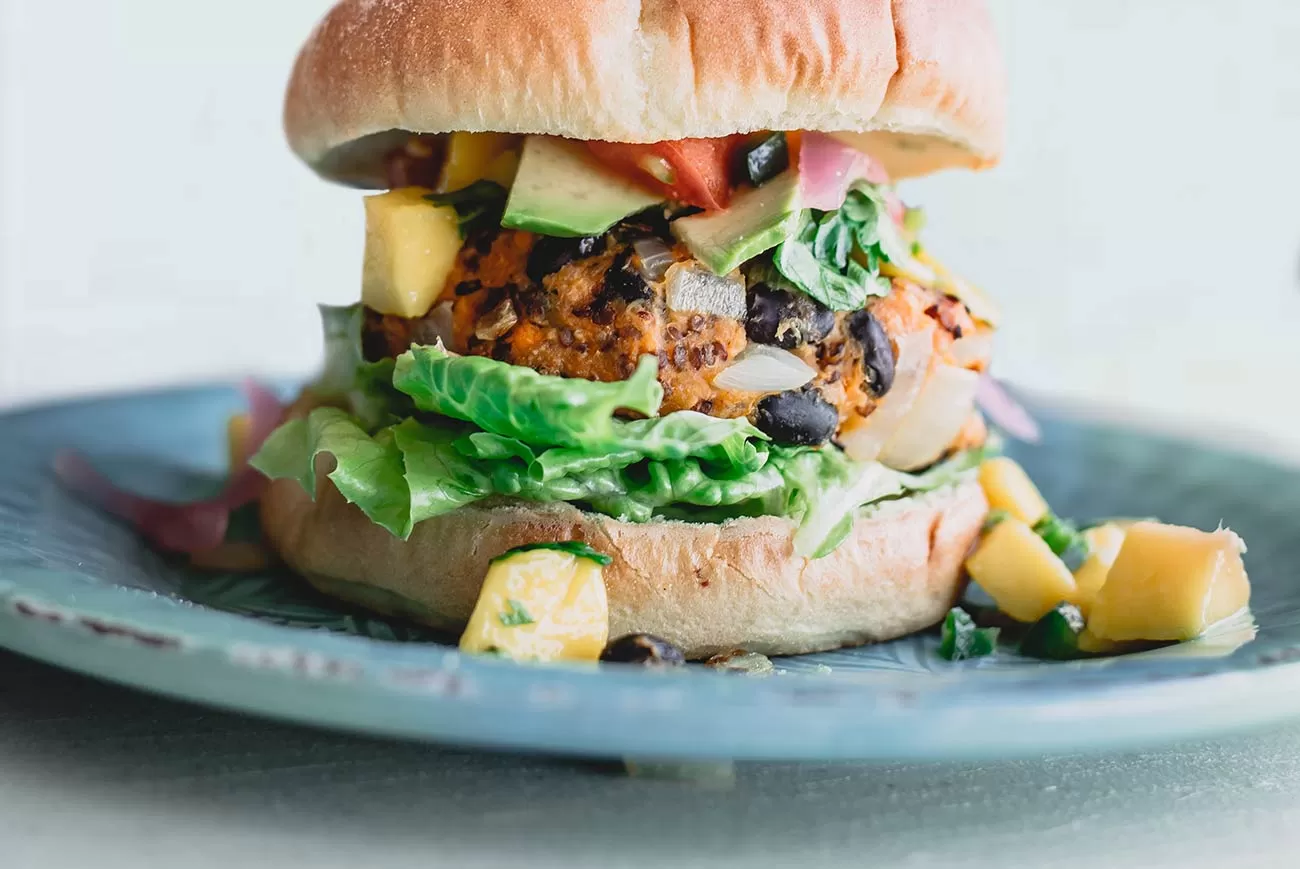 Close-up of a veggie burger with black beans, lettuce, avocado, tomato, and diced mango on a blue plate.