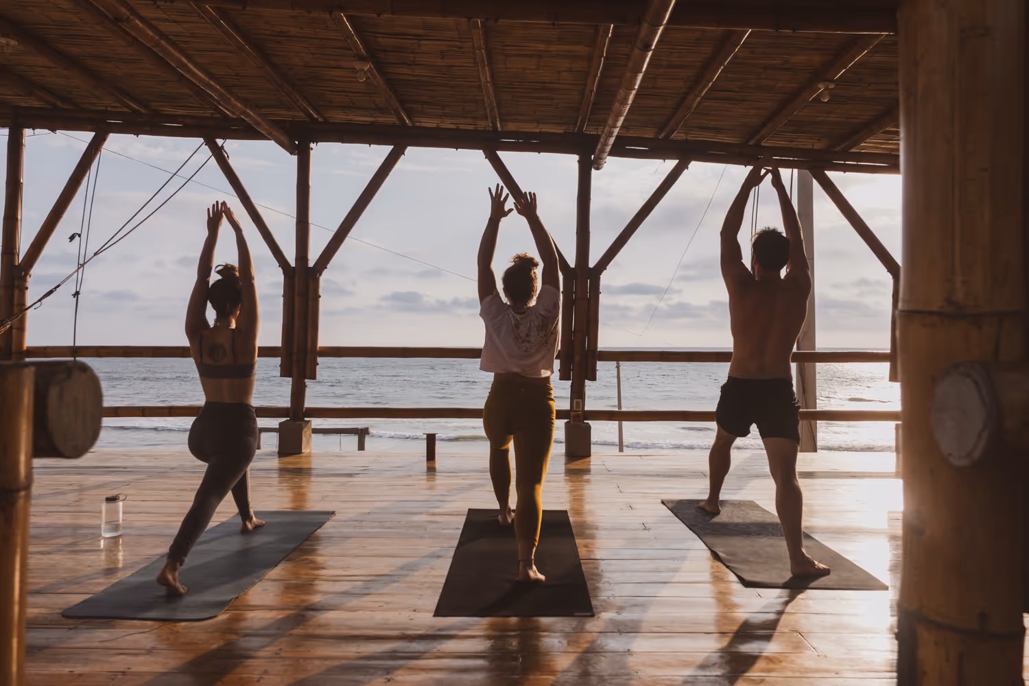 Three people doing yoga on mats under a bamboo shelter overlooking the ocean at sunset.