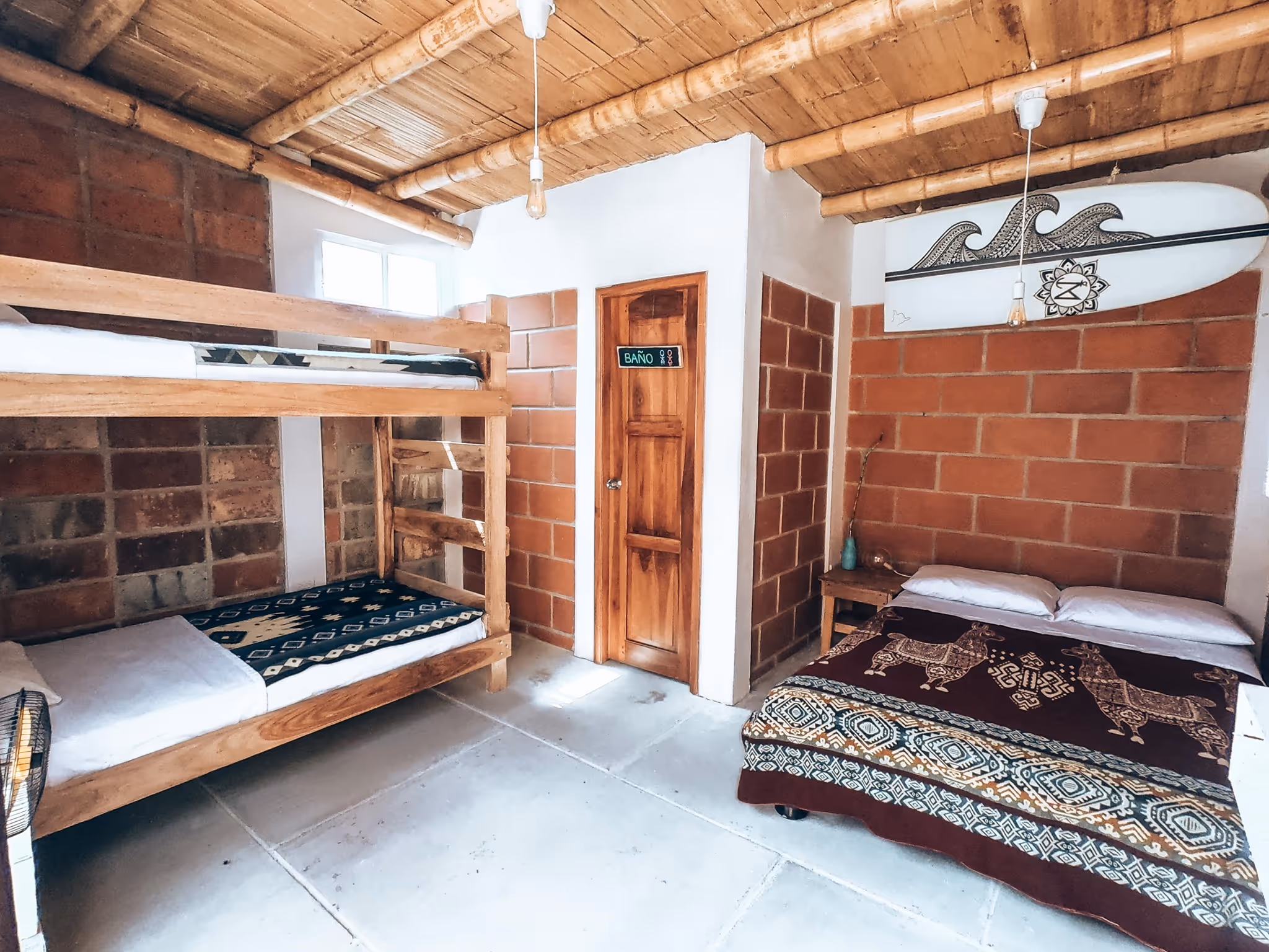 Rustic room with wooden bunk beds, a double bed with llama-patterned blanket, a wooden door labeled 'BAÑO', and a surfboard mounted on a brick wall.