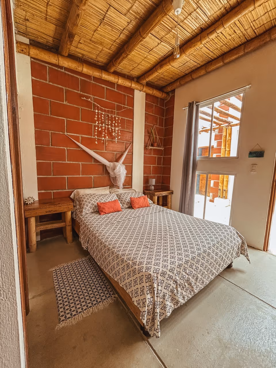 Cozy bedroom with a bed dressed in patterned bedding, two coral pillows, wooden nightstands, brick accent wall, bamboo ceiling, and window with curtain.