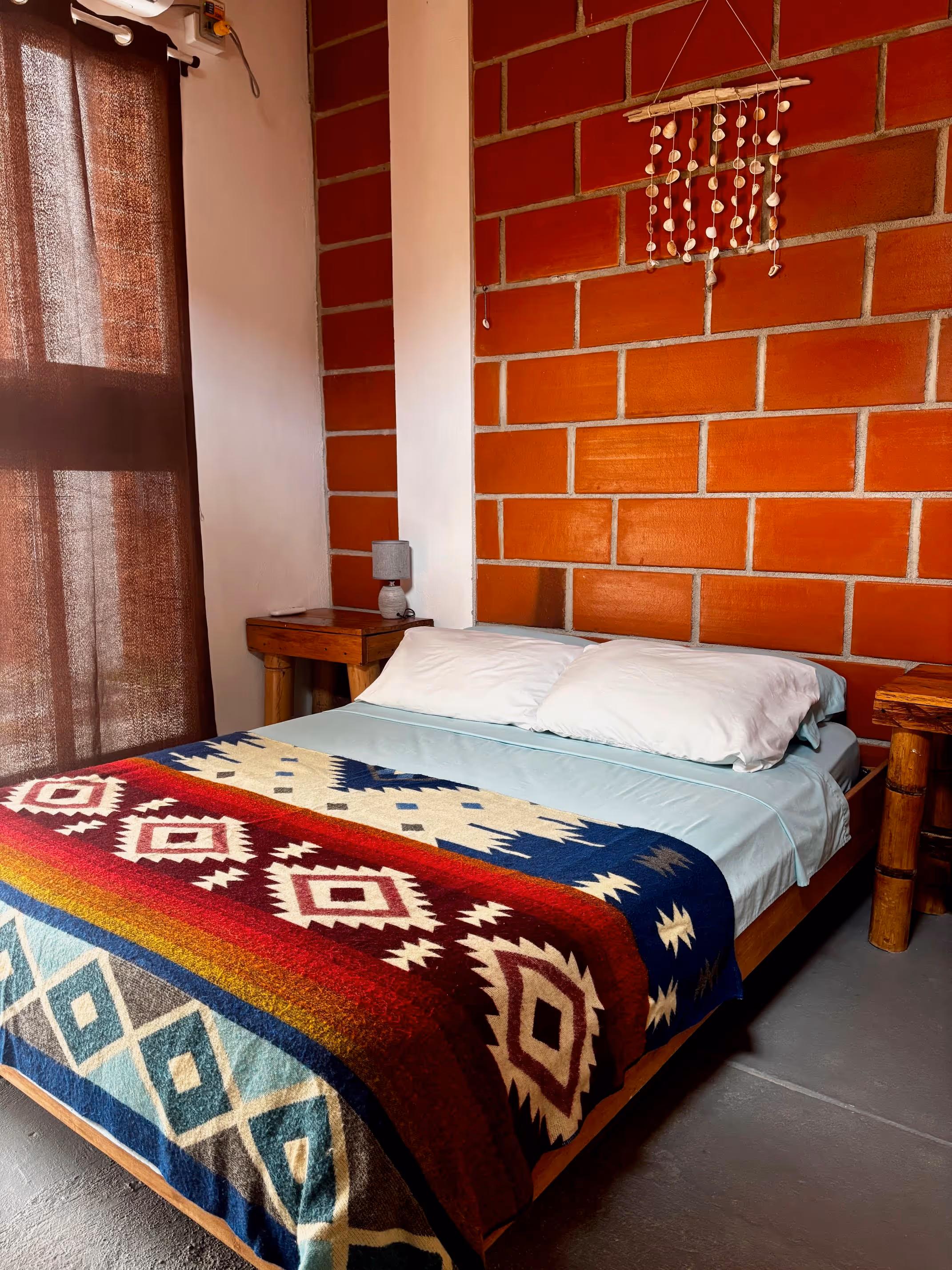 Cozy bedroom with a wooden bed, light blue sheets, white pillows, a colorful patterned blanket, a brick wall, and wooden furniture.