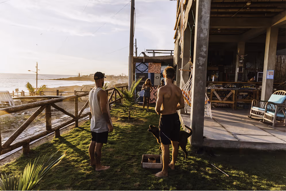 Two men standing barefoot on grass near a wooden fence by the ocean, with one man holding a dog and a woman in the background walking toward them.