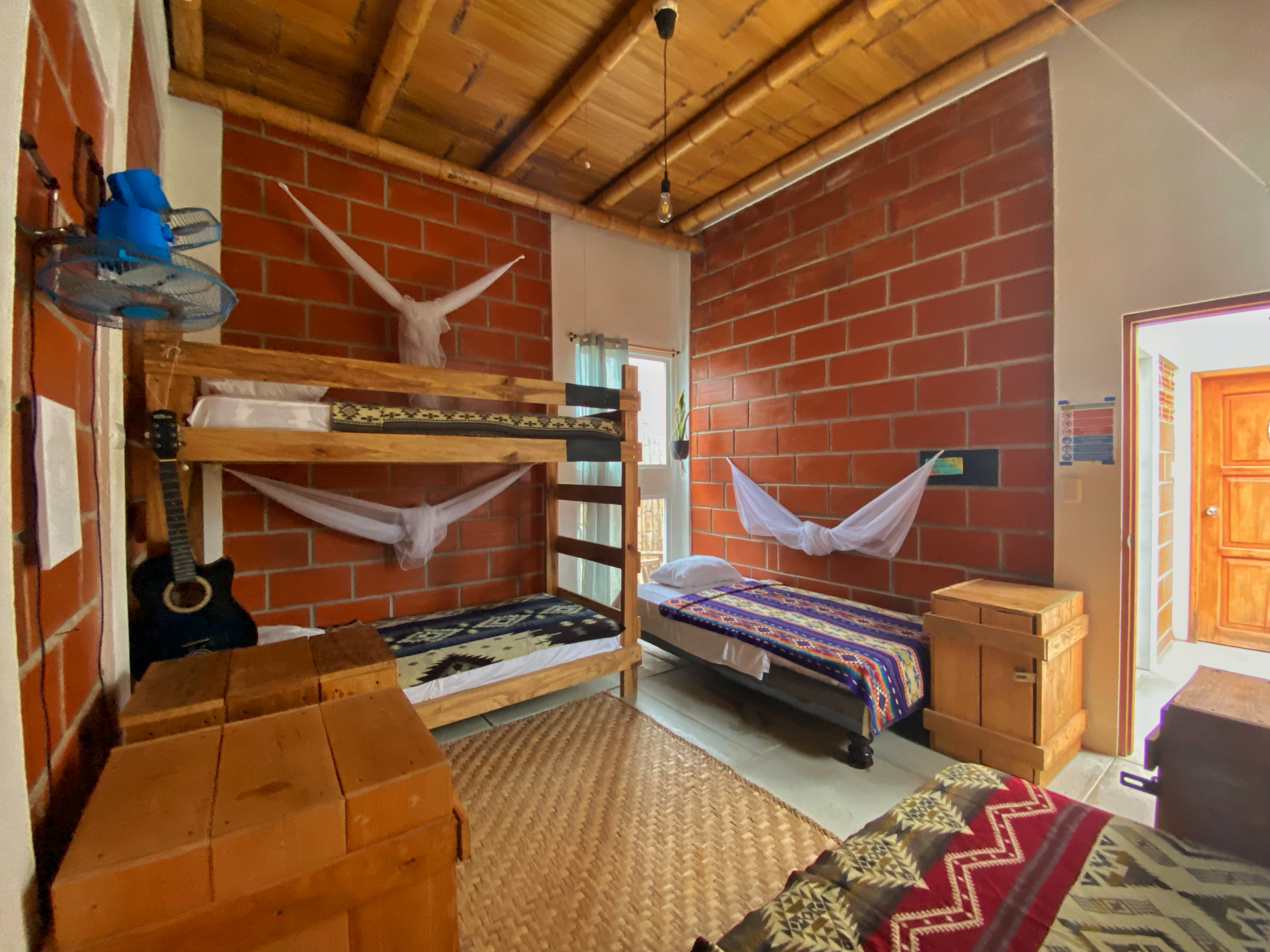 Rustic room with wooden bunk beds and single bed, colorful blankets, mosquito nets, woven rug, and a black guitar against a brick wall.