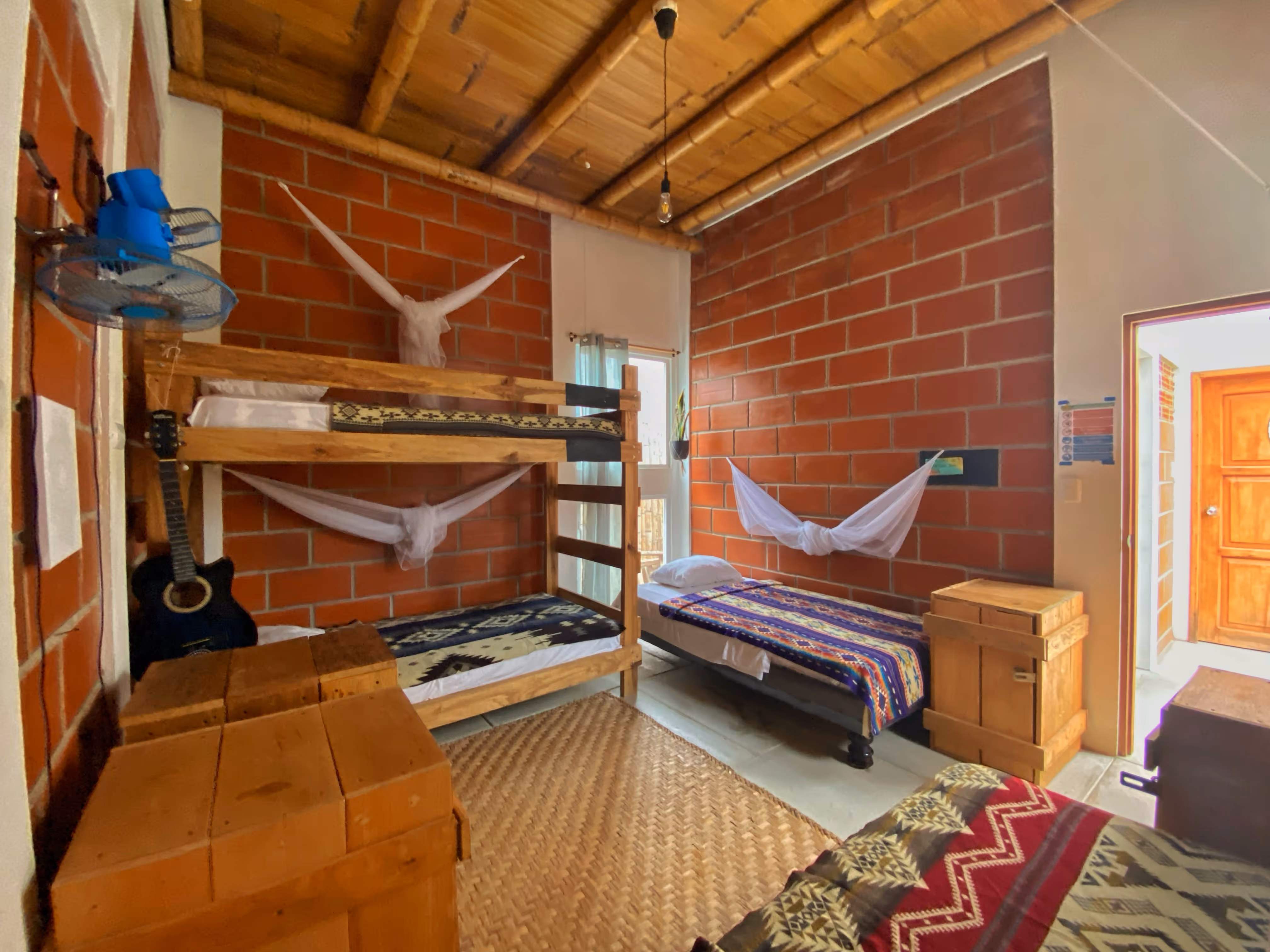 Rustic room with wooden bunk beds and single bed, colorful blankets, mosquito nets, woven rug, and a black guitar against a brick wall.