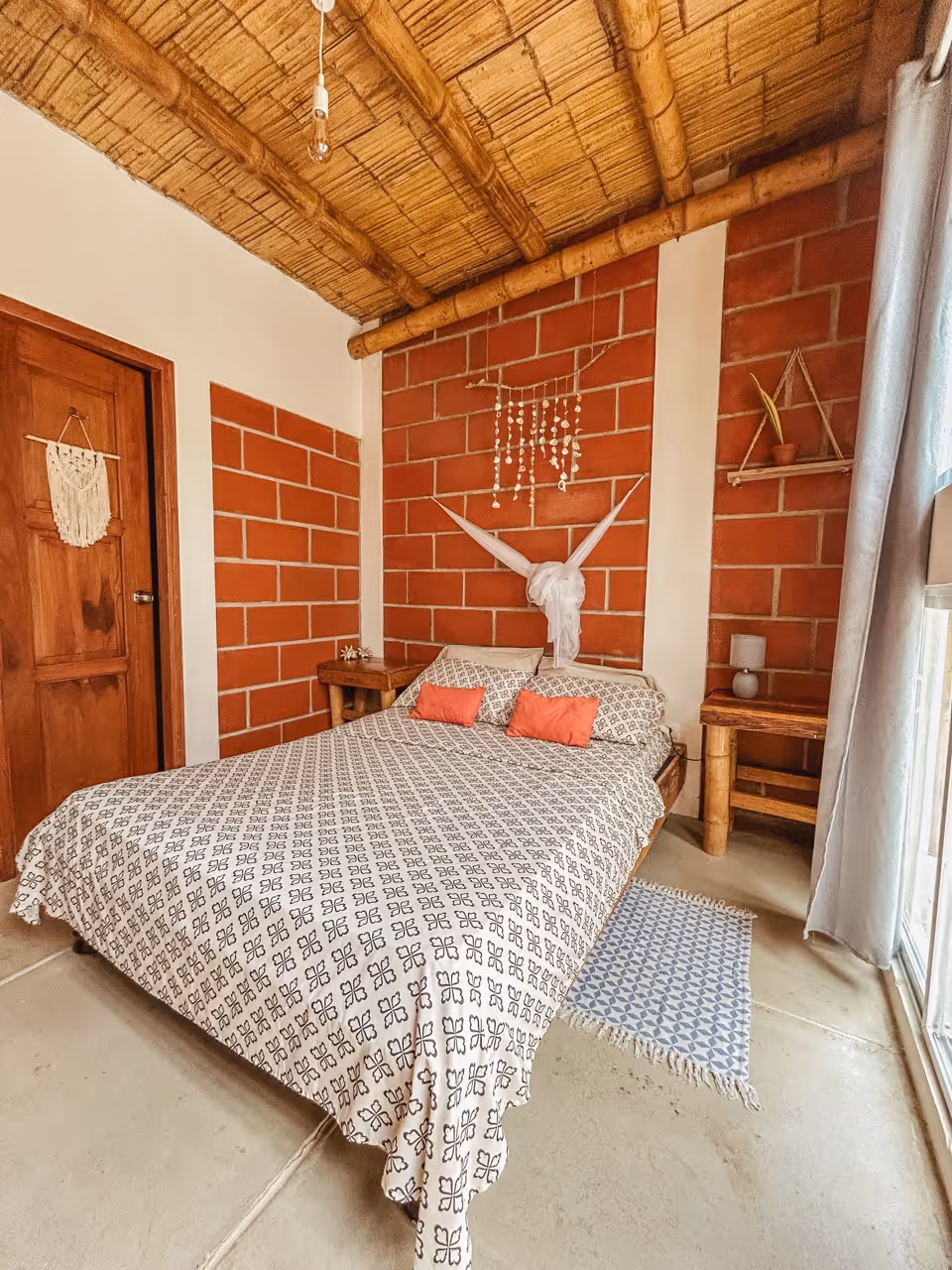 Cozy bedroom with patterned bedspread, coral accent pillows, brick walls, bamboo ceiling, and natural wood furniture.