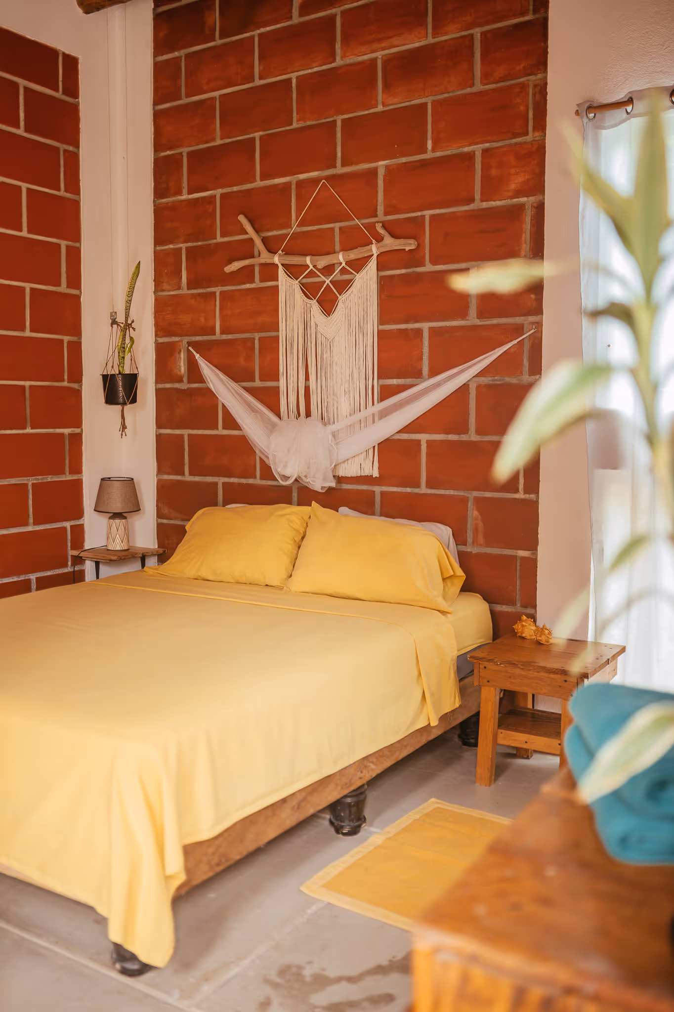 Bedroom with yellow bed sheets and pillows, a wooden nightstand, hanging plant, and macrame wall decor on a red brick wall.
