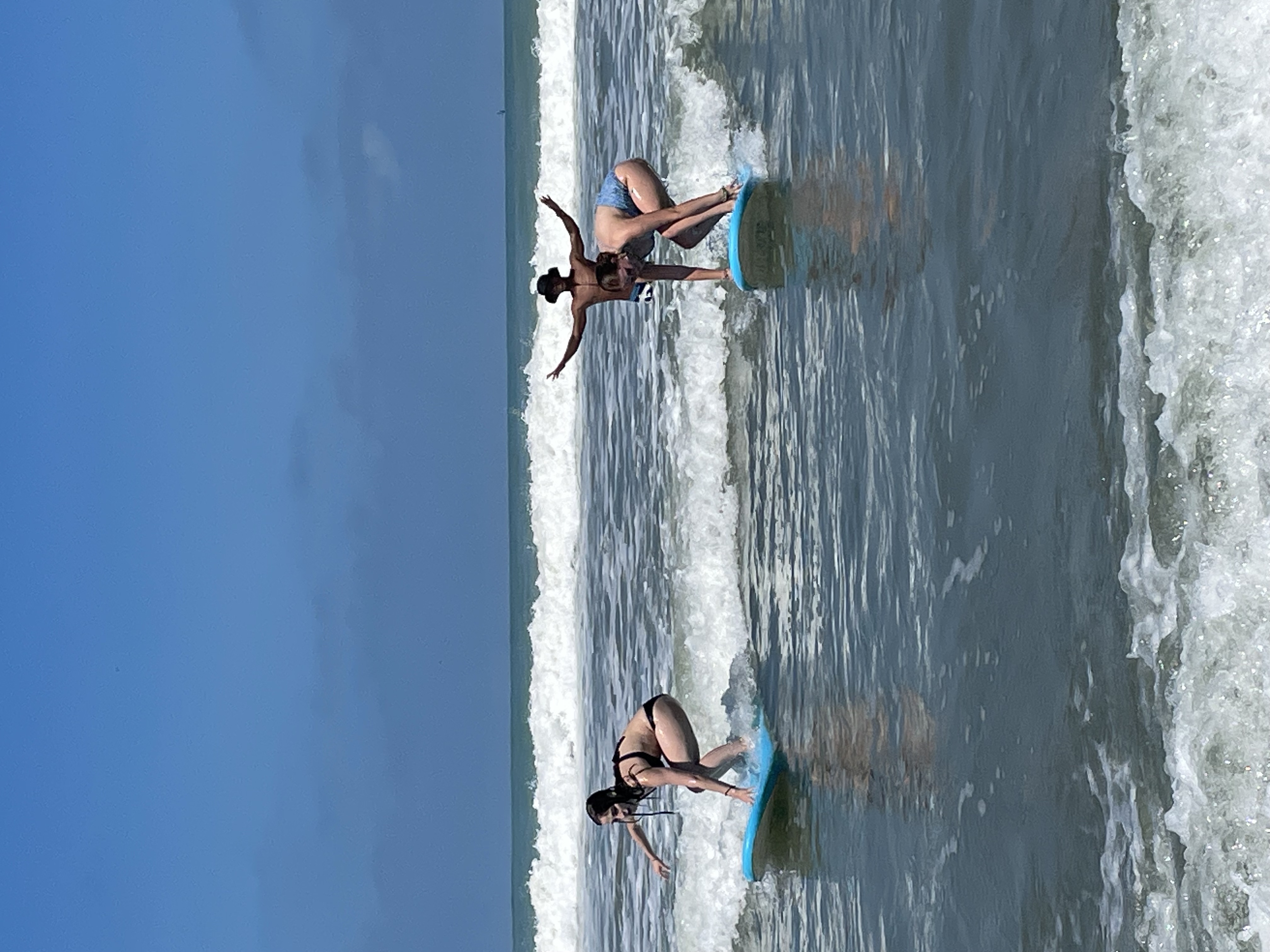 Two women learning to surf on small waves with a man standing in the ocean behind them raising his arms.