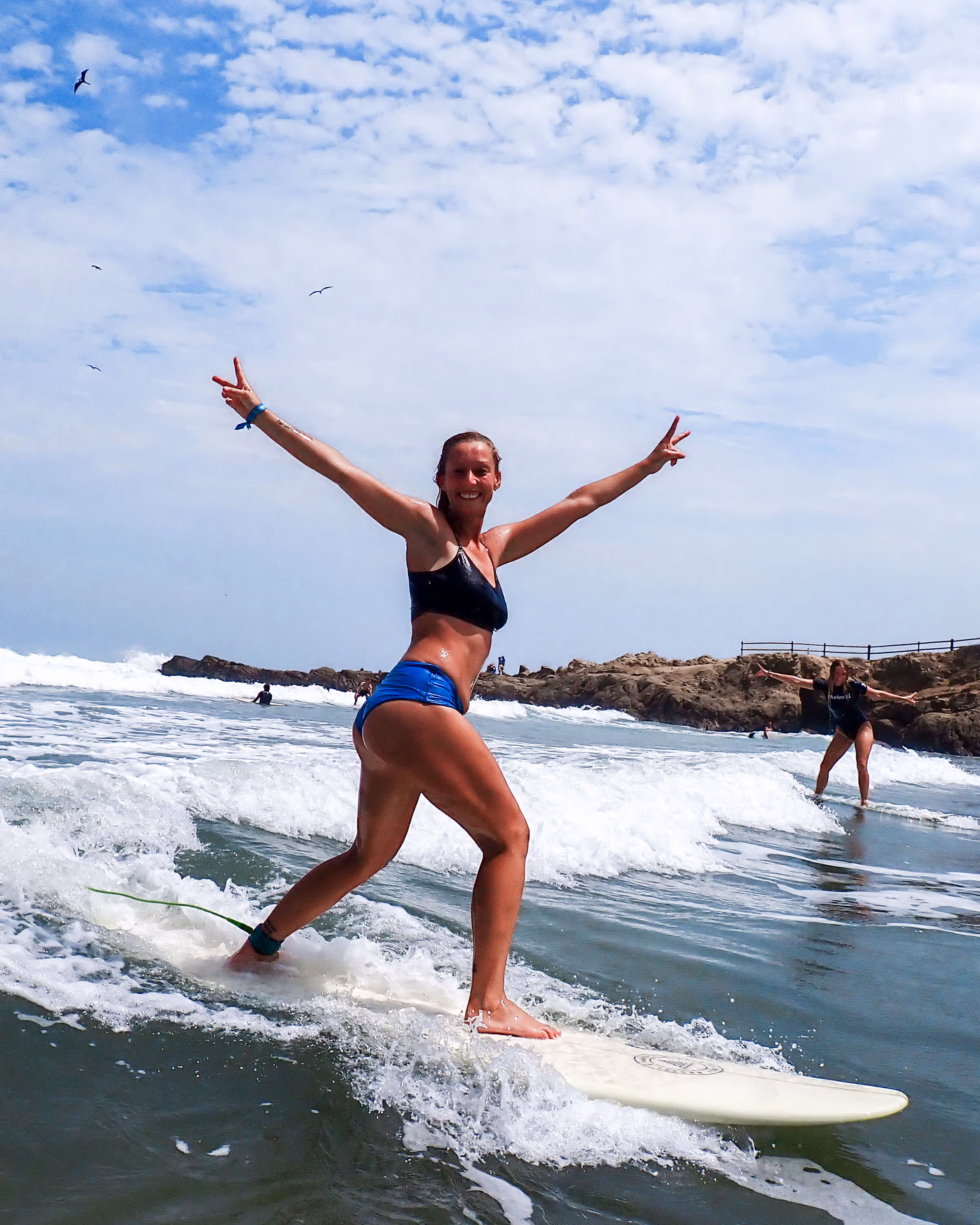 Smiling woman in a black top and blue shorts surfing on a wave with arms raised in peace signs under a partly cloudy sky.