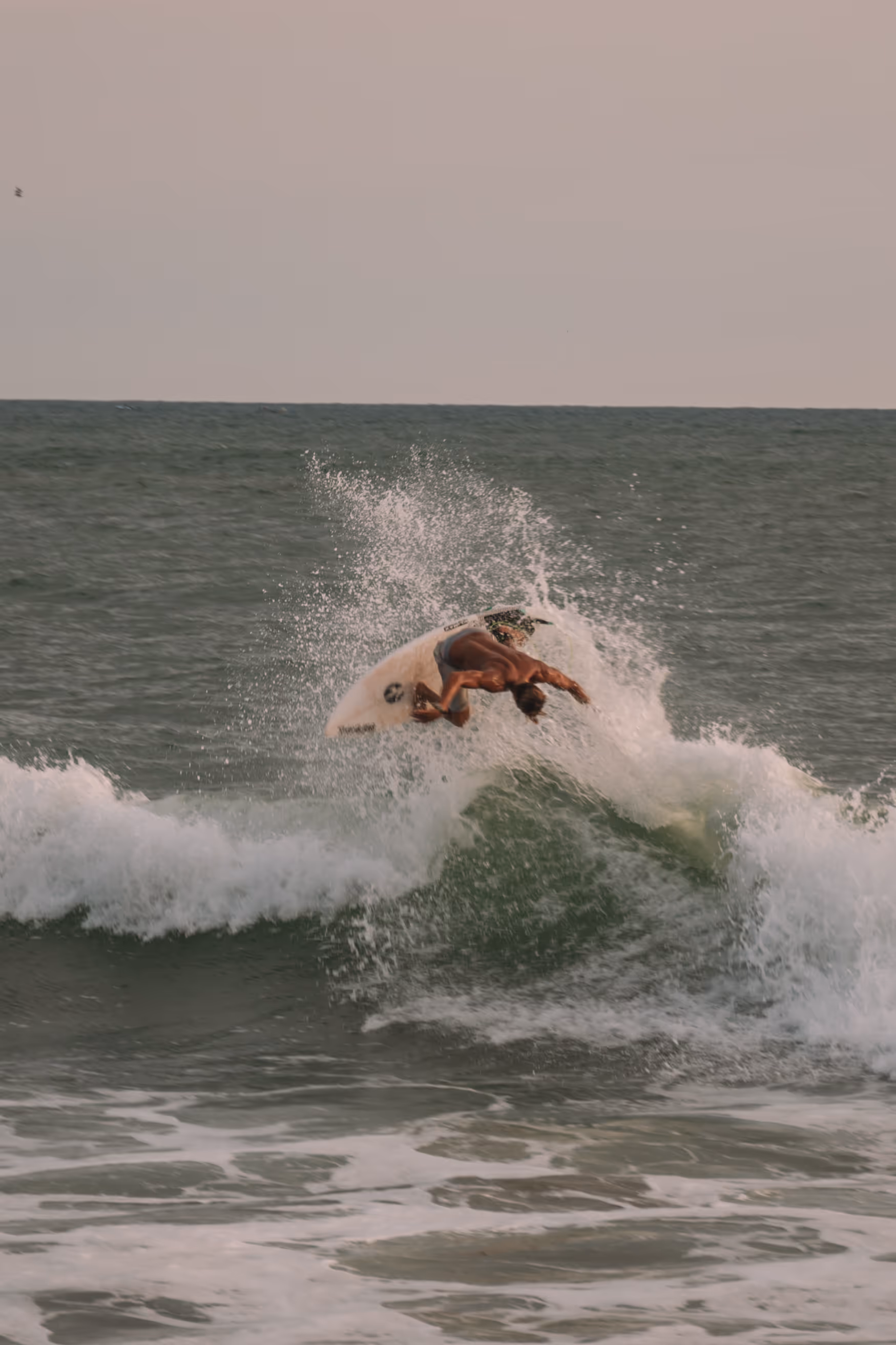 Surfer performing an aerial maneuver on a wave in the ocean under a hazy sky.