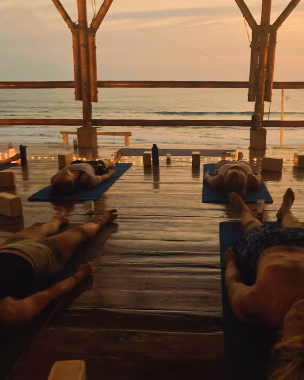Four people lying on yoga mats in a wooden pavilion overlooking the ocean at sunset, practicing relaxation.