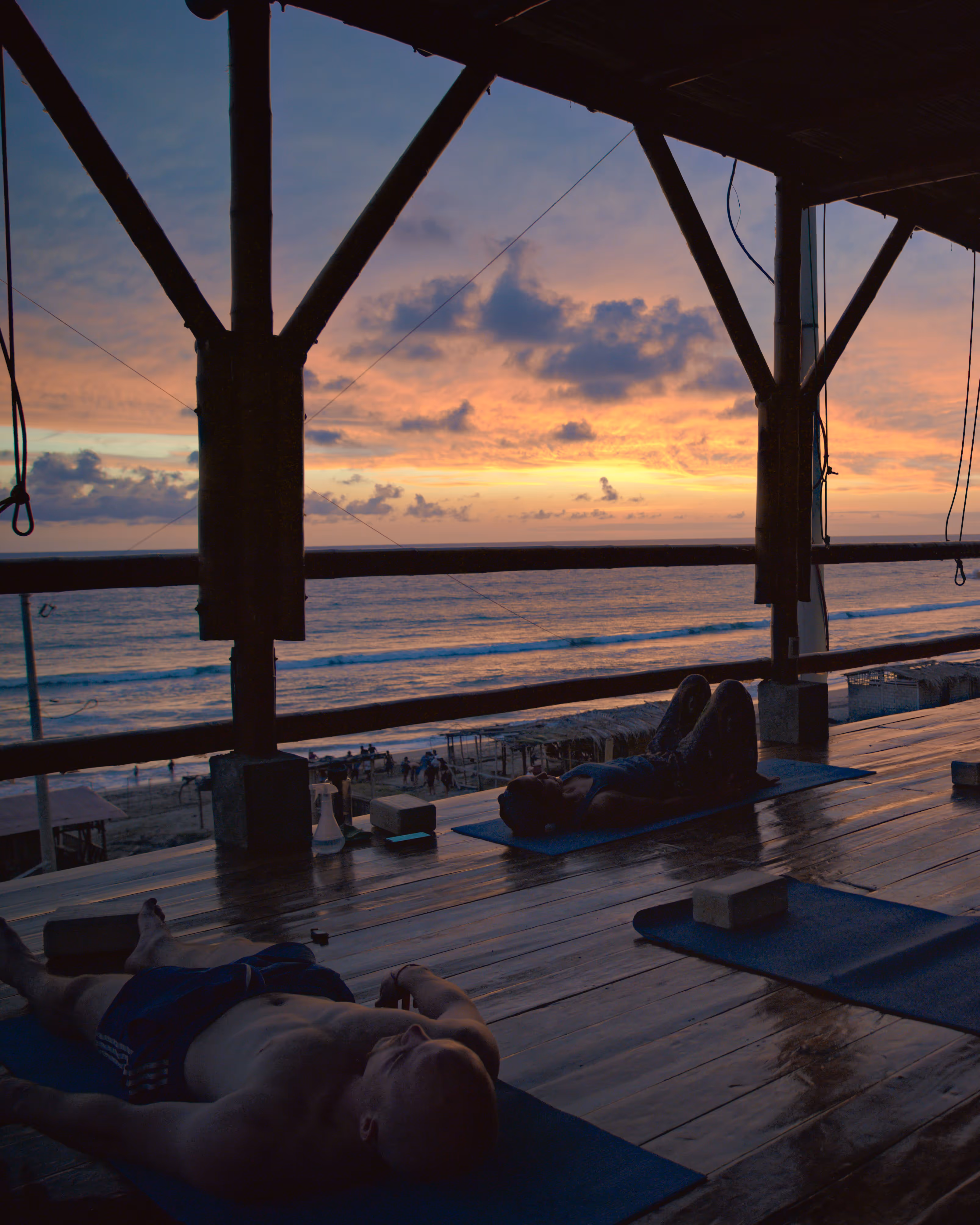 People relaxing on yoga mats inside a wooden pavilion overlooking the ocean during a colorful sunset.