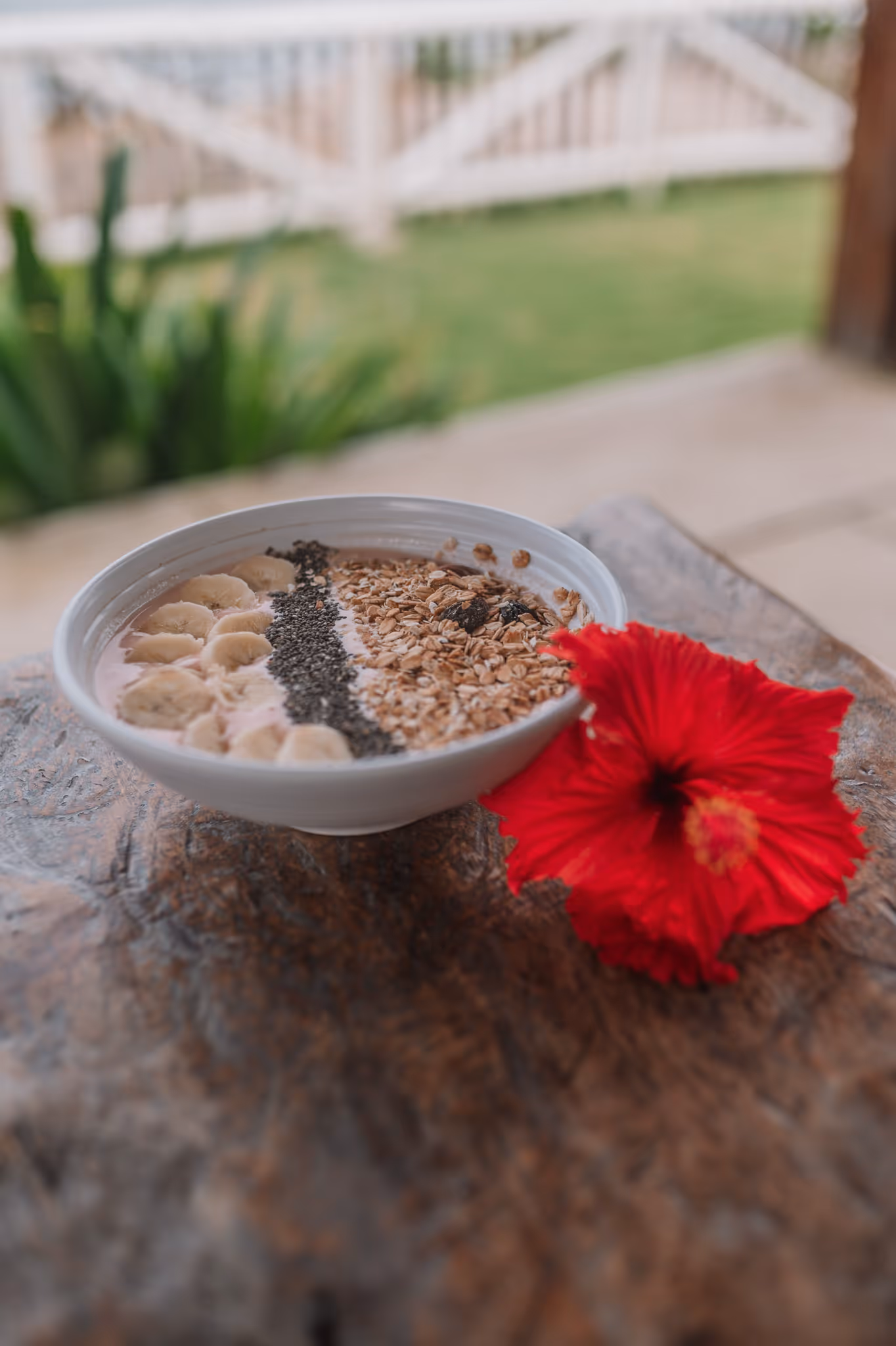 Smoothie bowl topped with banana slices, chia seeds, and granola with a red hibiscus flower beside it on a wooden surface.