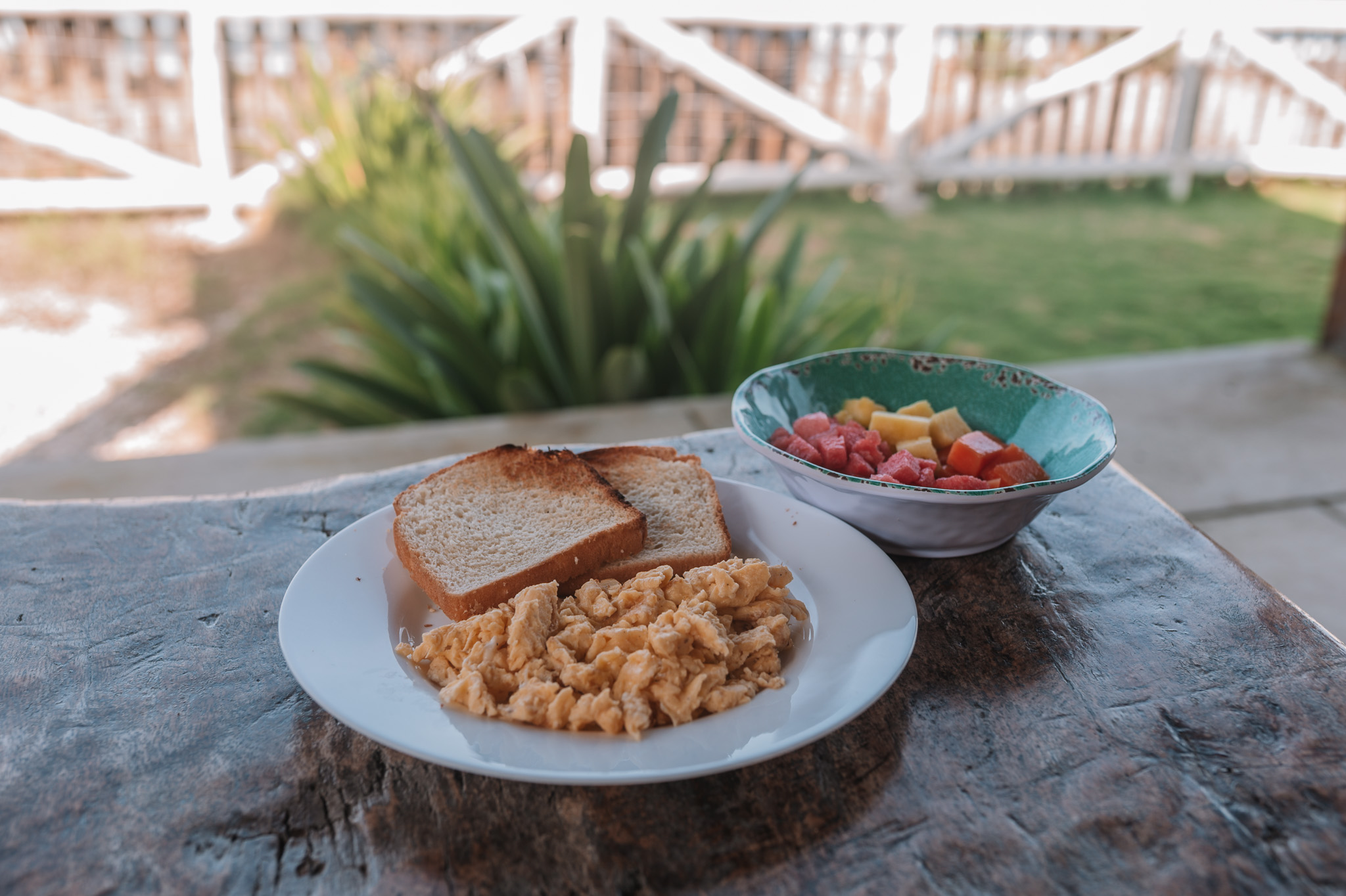 Plate with scrambled eggs and two slices of toasted bread next to a bowl of colorful fruit salad on a wooden table outdoors.