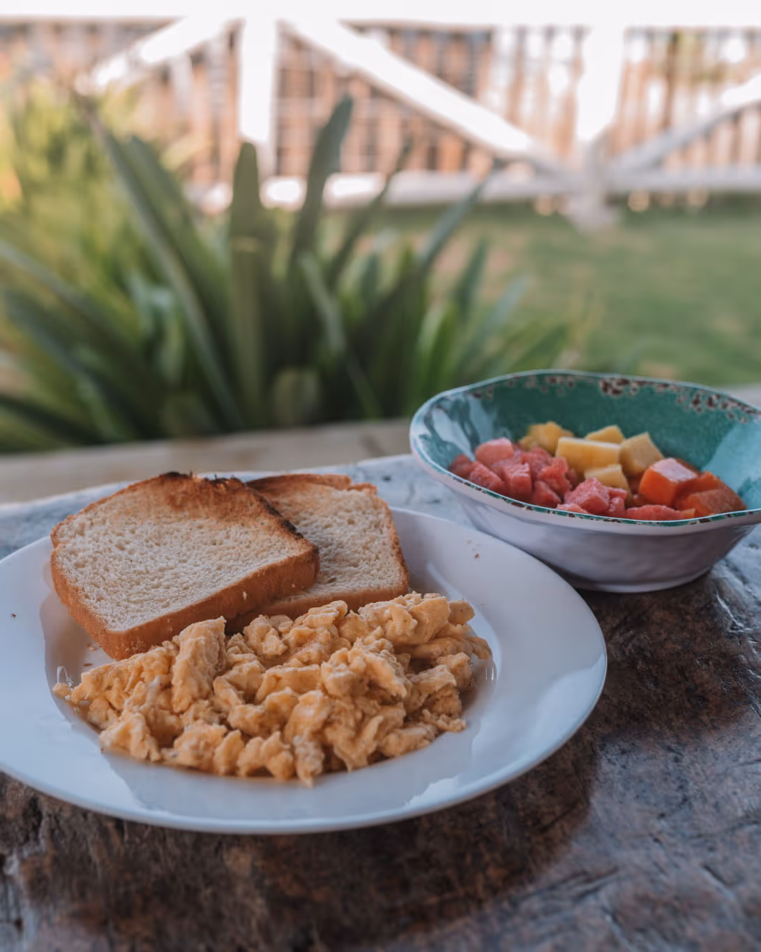 Plate with scrambled eggs and two slices of toasted bread next to a bowl of mixed fresh fruit on a wooden table outdoors.