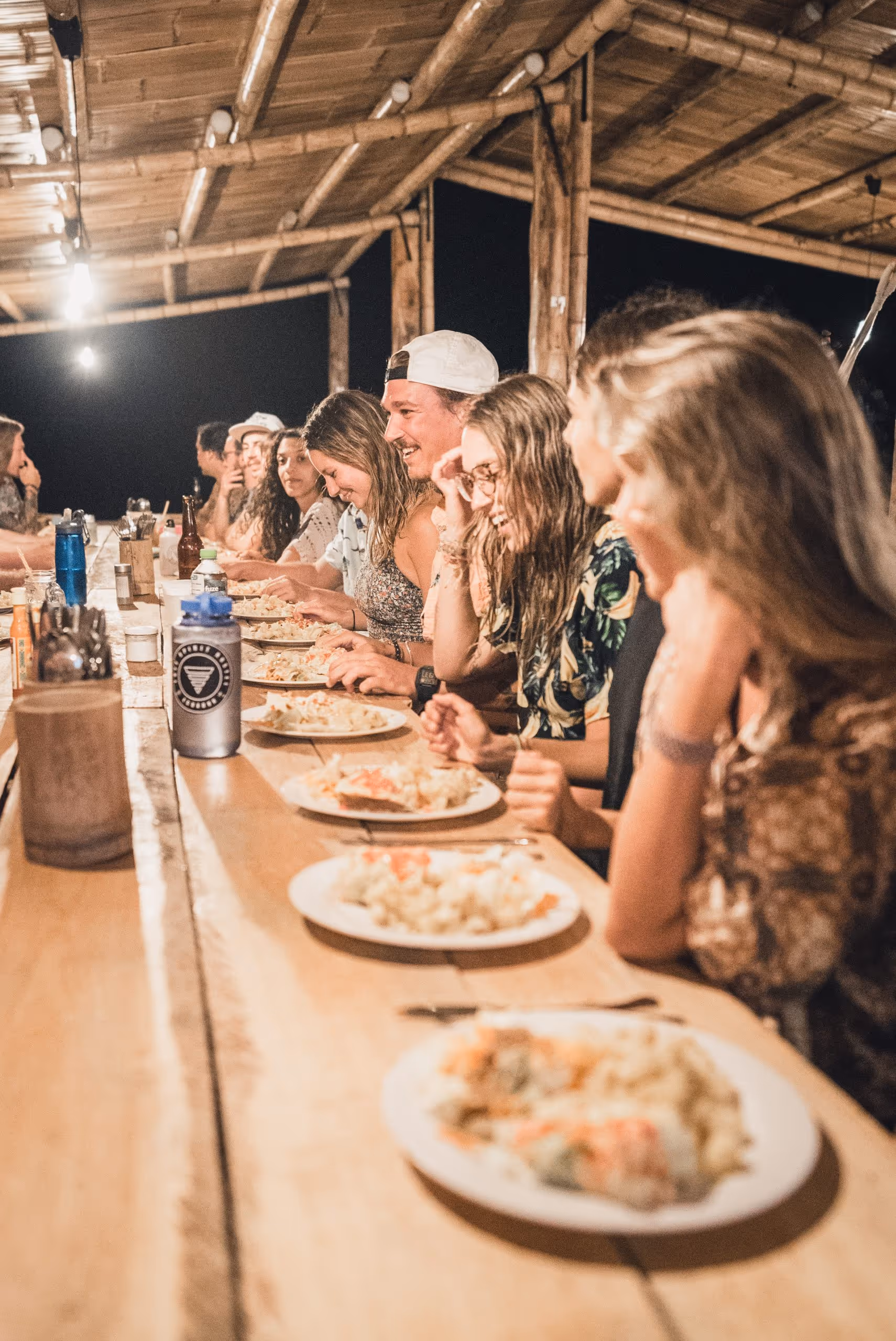 Group of people sitting at a long wooden table enjoying a meal together under warm lighting in a bamboo-structured outdoor setting at night.