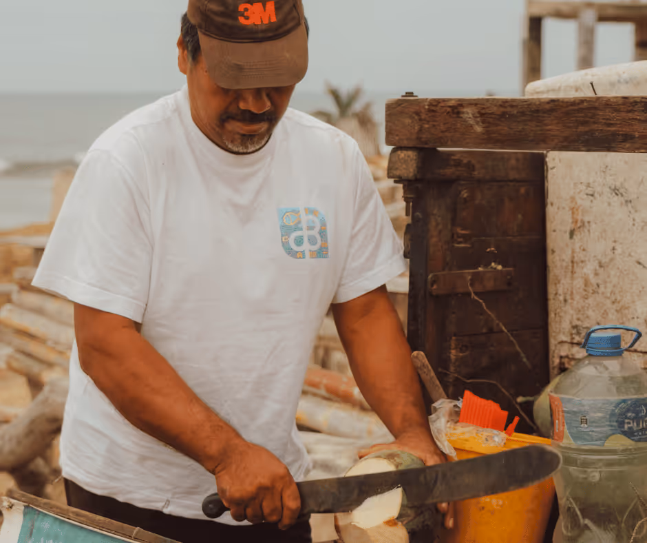 Man wearing a brown 3M cap and white t-shirt cutting a coconut with a large machete outdoors.
