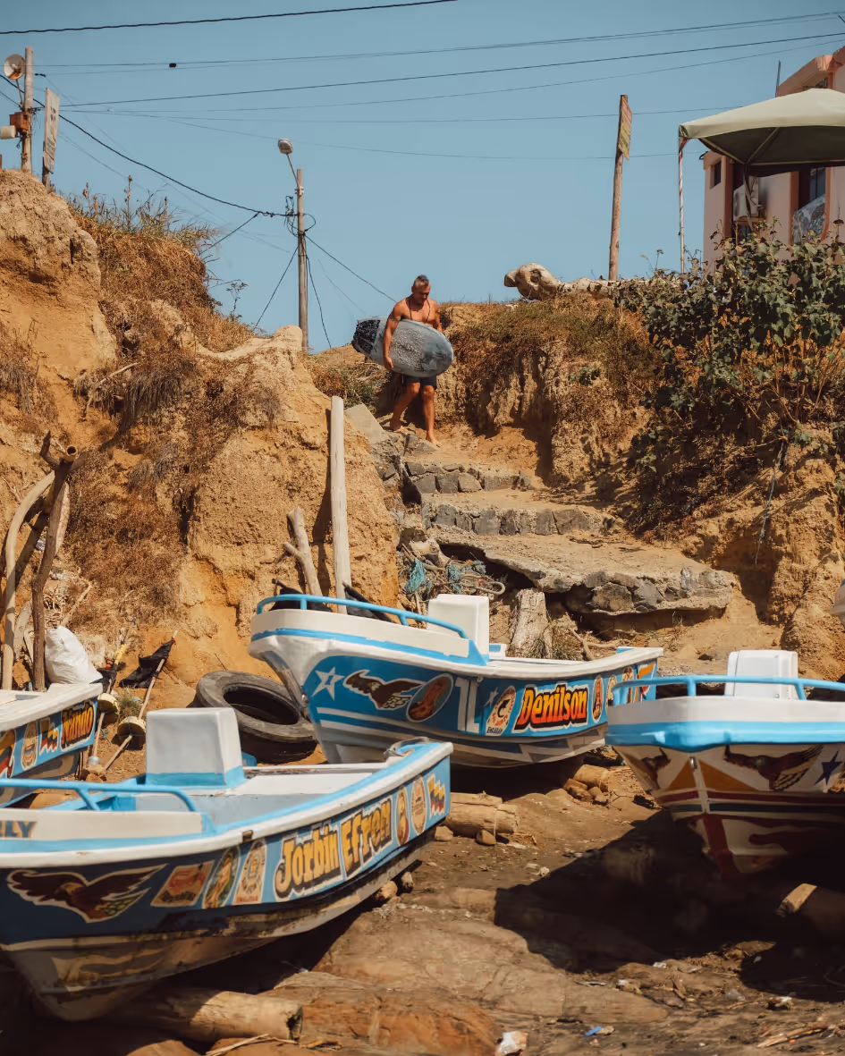 Man carrying a surfboard down stone steps near painted fishing boats on a rocky shore.