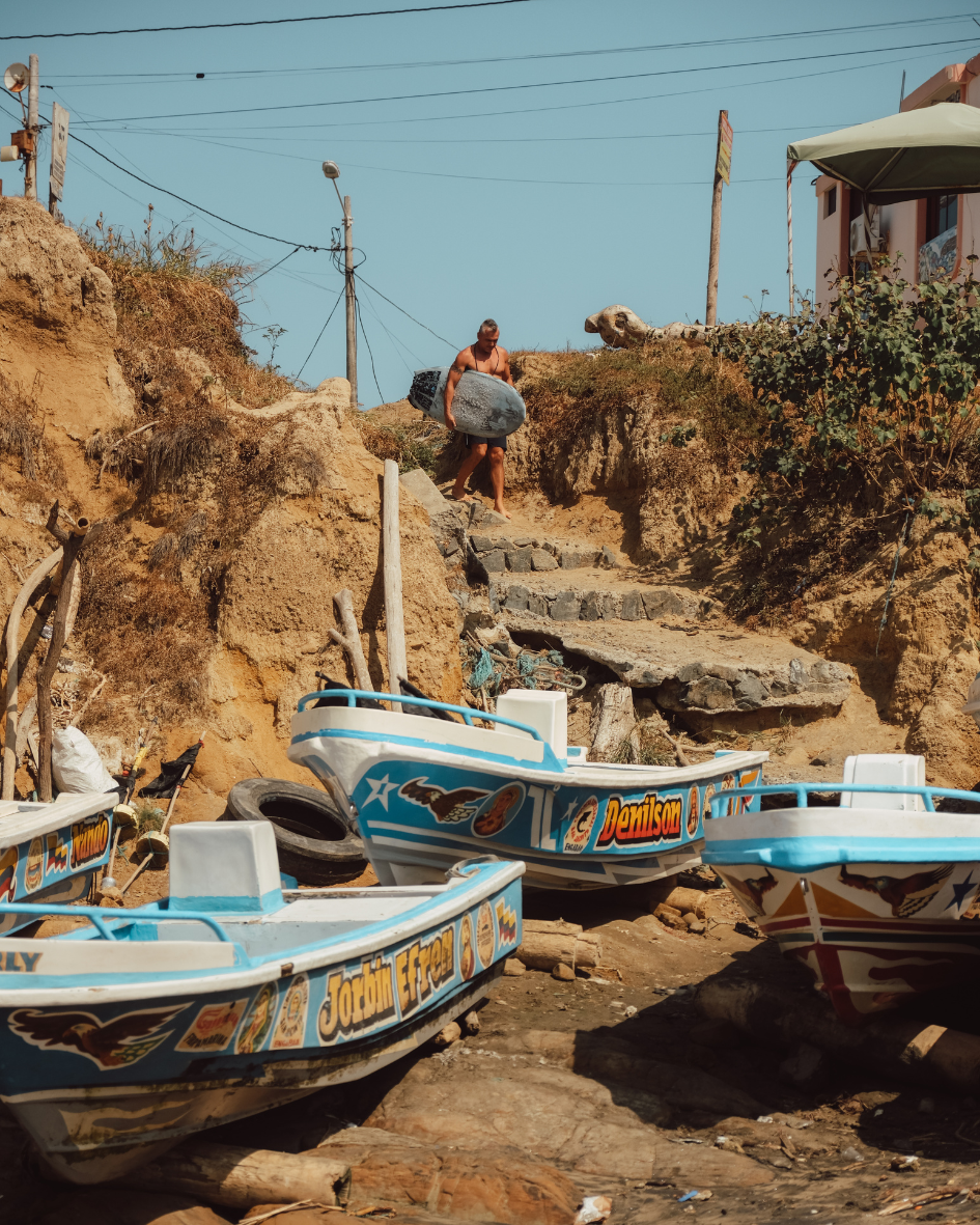 Man carrying a surfboard down stone steps near painted fishing boats on a rocky shore.