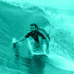 Man surfing on a wave with a beard and dark shirt.