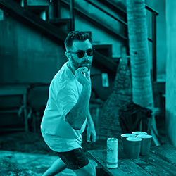 Man wearing sunglasses and a white shirt playing beer pong indoors.