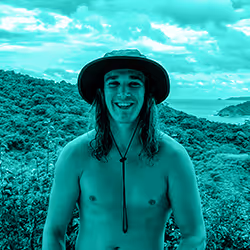 Smiling shirtless young man wearing a wide-brimmed hat standing outdoors with a forest and ocean background under a cloudy sky.