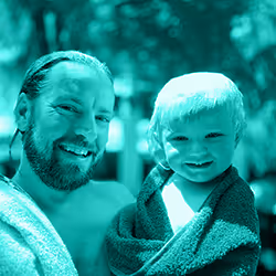Smiling man with wet hair and beard holding a happy young child wrapped in a towel outdoors.
