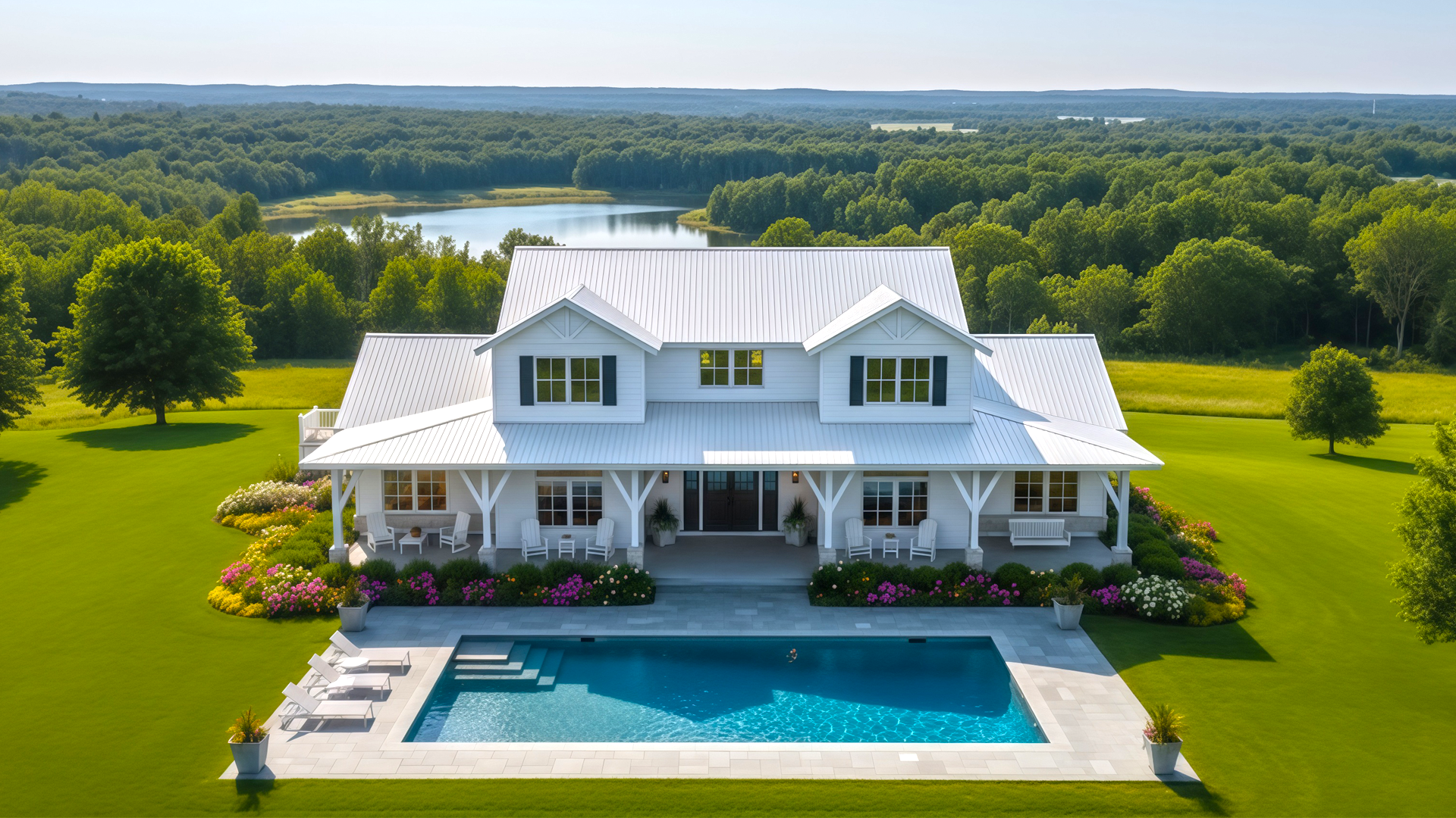 White two-story farmhouse with black shutters, a large porch, surrounded by colorful flower beds, a rectangular swimming pool with lounge chairs, and expansive green lawn with trees and a distant lake.