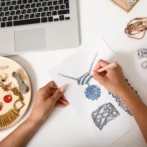 Hands sketching jewelry designs on paper near a laptop and jewelry display on a white table.