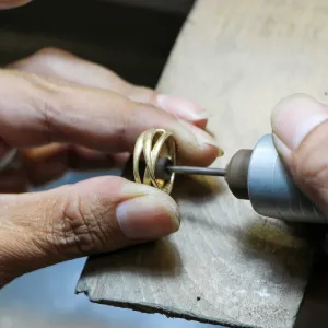 Hands polishing or drilling a gold ring on a workbench with a rotary tool.