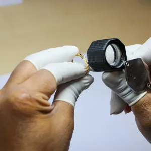 Hands with white gloves inspecting a gold ring using a jeweler's loupe.