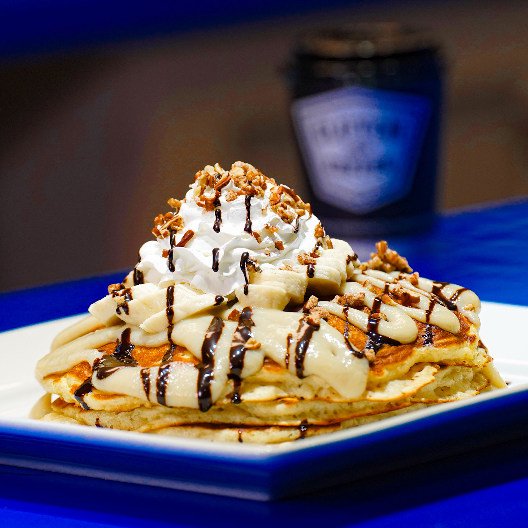 Three buttermilk pancakes topped with sliced banana, banana puree, candied pecans, whip cream, powdered and chocolate drizzle